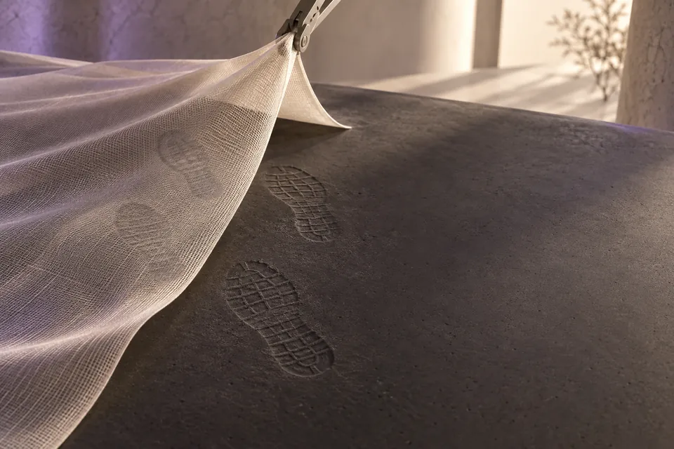 A translucent technical fabric lifts to reveal footprints already impressed in a charcoal surface.